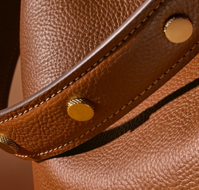 Eine Grafik mit einem „F“. Das braune Lederband mit goldfarbenen Nieten, die unsere dezente Signature-Knurling-Struktur zeigen.