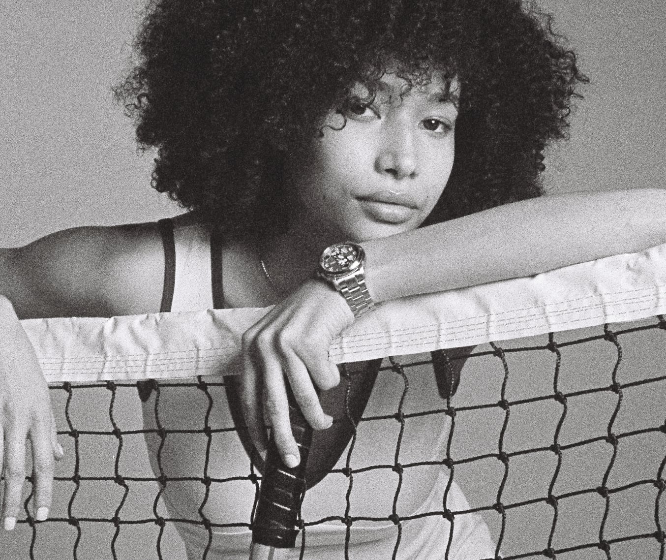 A black and white image of a woman casually leaning over a tennis net, holding a tennis racket. She's wearing a white tennis dress and our limited-edition Disney Mickey Mouse Tennis Watch.