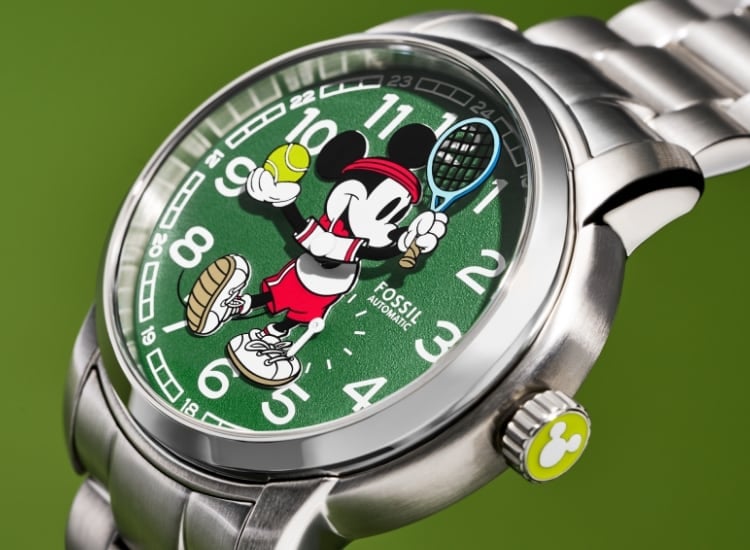 Notre montre Mickey Mouse Disney Tennis en série limitée. Sur fond de cadran vert texturé inspiré de la surface d’un court de tennis, Mickey porte un polo blanc et des baskets, un bandeau rouge et un short rouge. Il tient une raquette bleue dans une main et une balle de tennis dans l’autre. Ses bras bougent, indiquant les heures et les minutes.